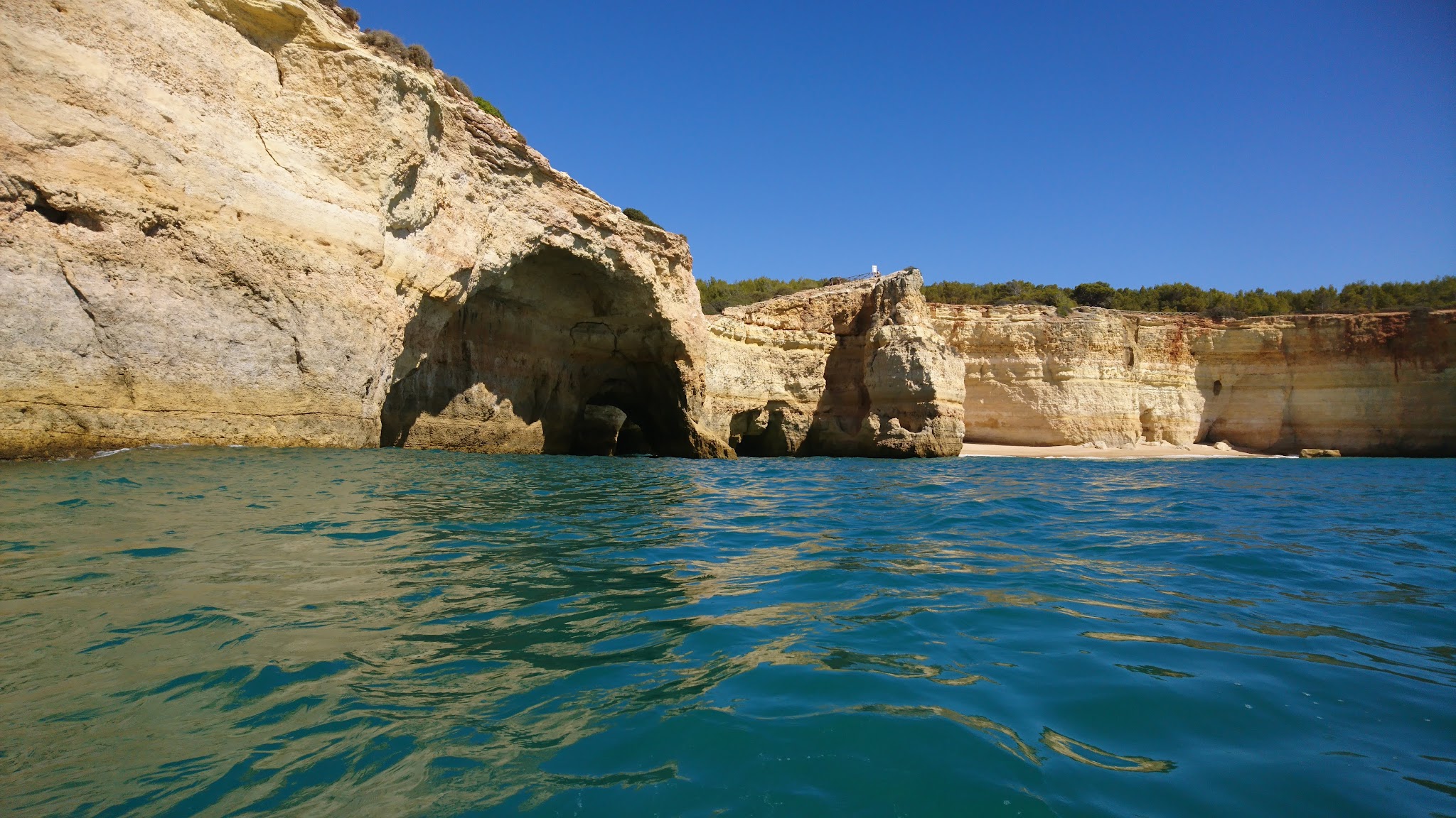 Helle Kalksteinklippe mit großer Meereshöhle, davor türkisblaues Wasser unter wolkenlosem Himmel.