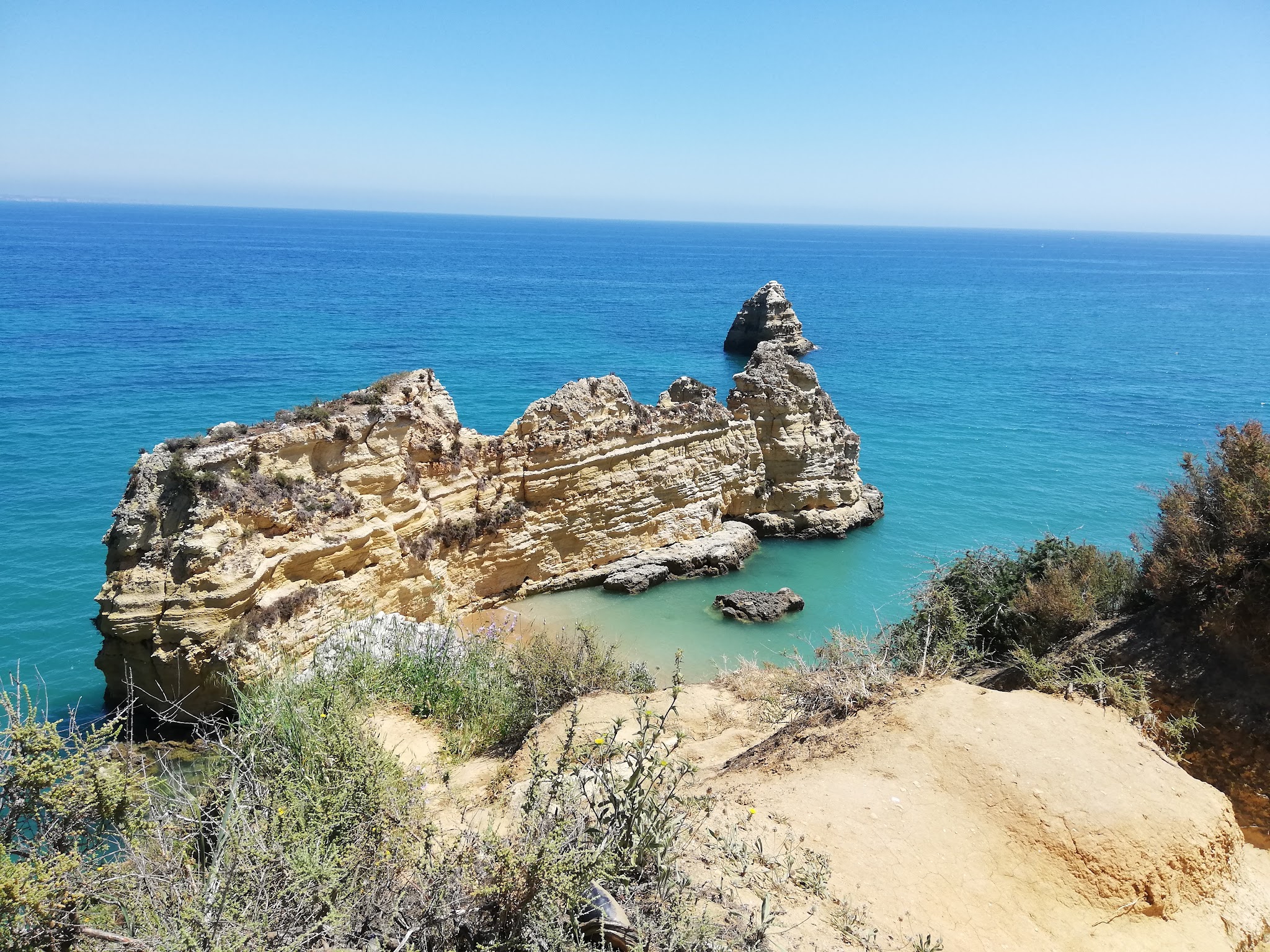 Gelblich zerklüftete Felsklippe ragt ins türkisblaues Meer, umgeben von klarem Himmel und spärlicher Küstenvegetation.