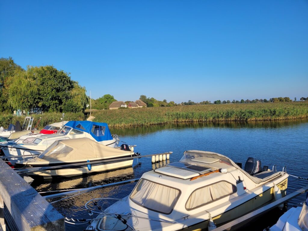 Mehrere kleine Motorboote liegen an Stegen auf ruhigem Fluss, gegenüber Schilf und Bäume unter klarem blauem Himmel.