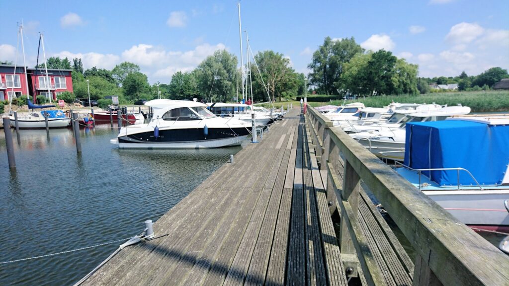 Holzsteg mit festgemachten Motor- und Segelbooten in ruhigem Yachthafen bei Sonne und blauem Himmel.