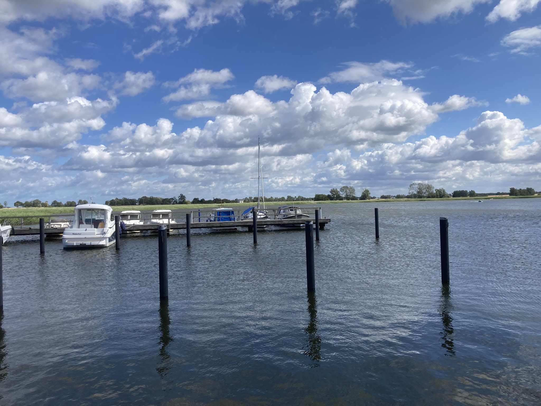 Kleine Anlegestelle mit mehreren Motorbooten, leeren Pfählen, ruhigem Wasser und Wolkenhimmel im Hintergrund.