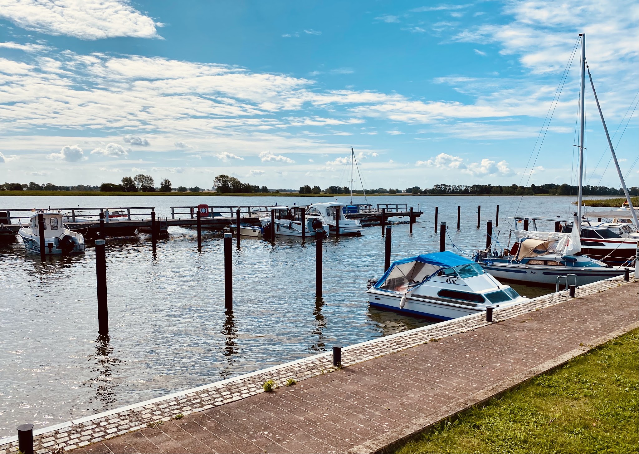 Kleiner Yachthafen: Mehrere Motor- und Segelboote liegen an Stegen, ruhiges Wasser unter blauem Himmel mit Wolken.
