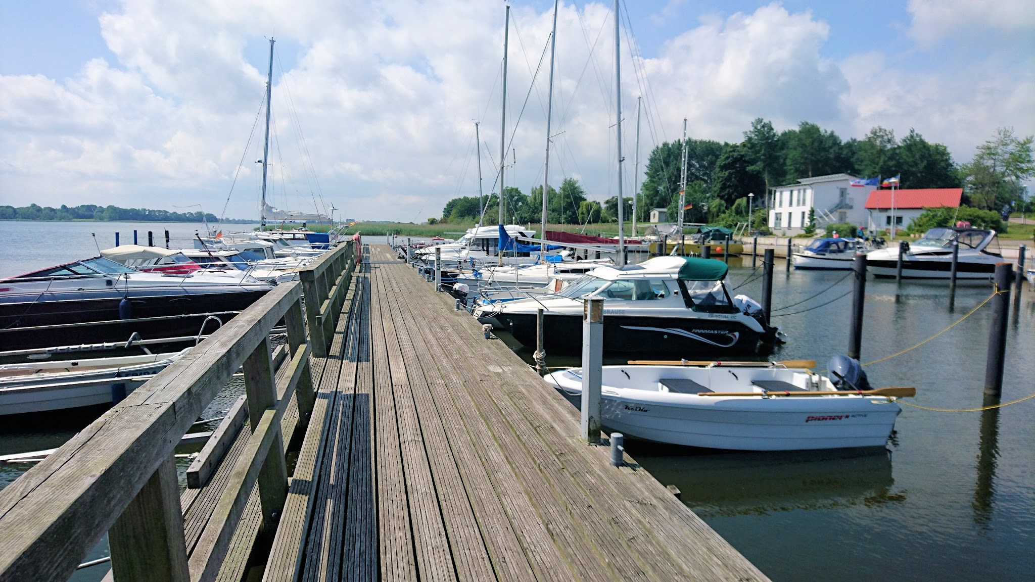 Langer Holzsteg mit angelegten Segel- und Motorbooten in sonnigem Yachthafen, ruhiges Wasser und blauer Himmel.