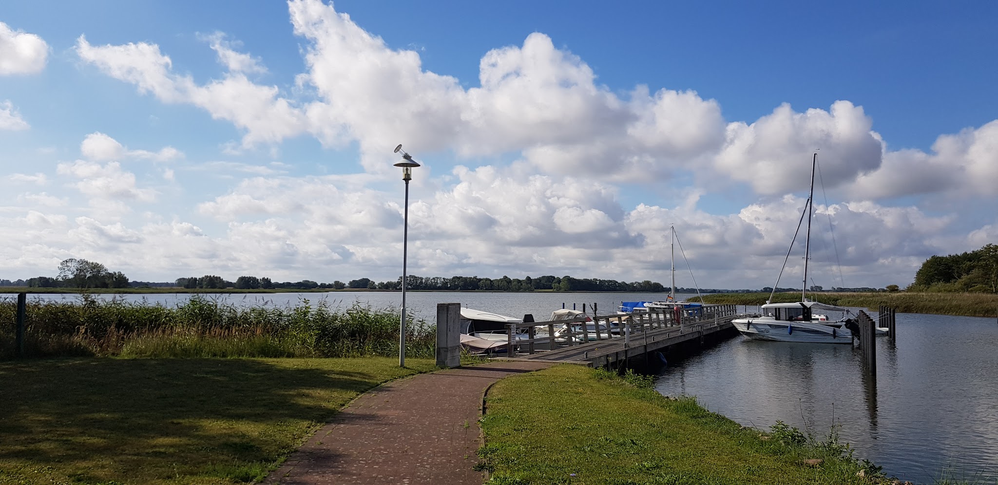 Grasweg führt zu Steg mit Segel- und Motorbooten; Möwe auf Laterne, ruhiger See unter blauem Himmel mit Wolken.