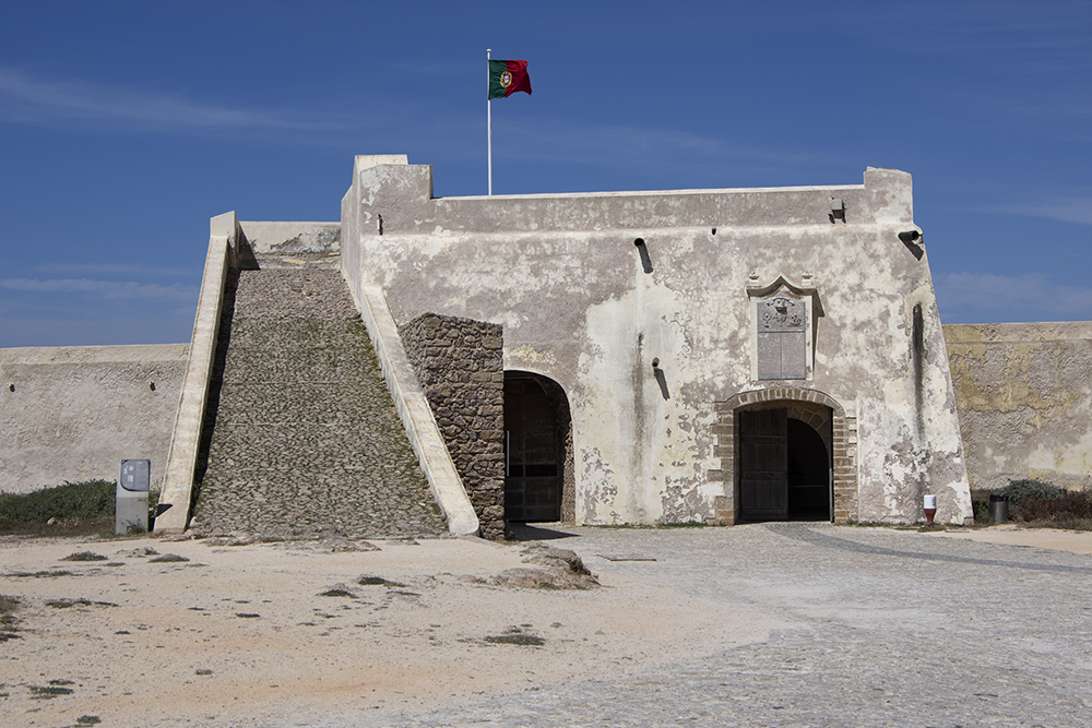 Altes Küstenfort mit breiter Steinrampe und zwei Torbögen; portugiesische Flagge weht unter klarem Himmel.