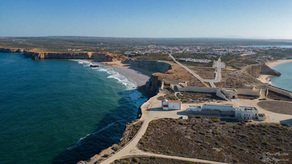 Luftaufnahme: historische Festung auf Felsklippe, darunter türkisblaues Meer und Strand, dahinter Küstenort.