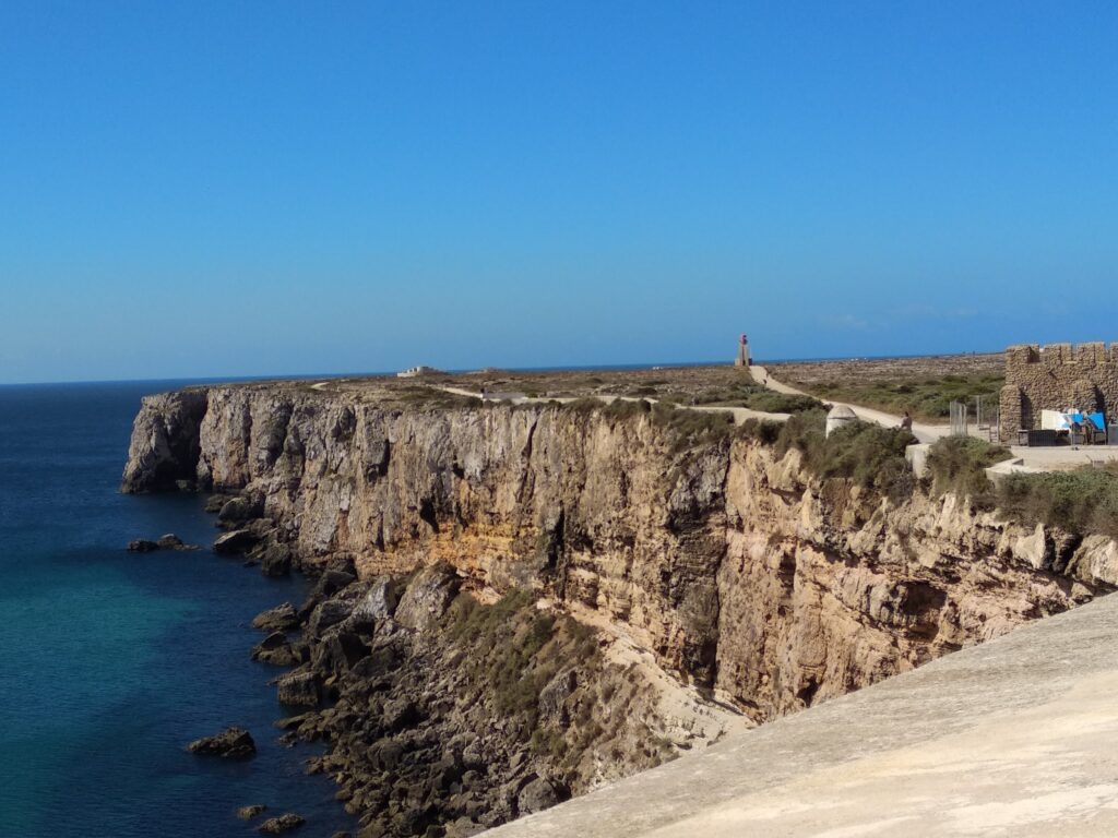 Steile, zerklüftete Küstenklippe mit tiefblauem Atlantik; in der Ferne ein kleiner Leuchtturm unter klarem Himmel.