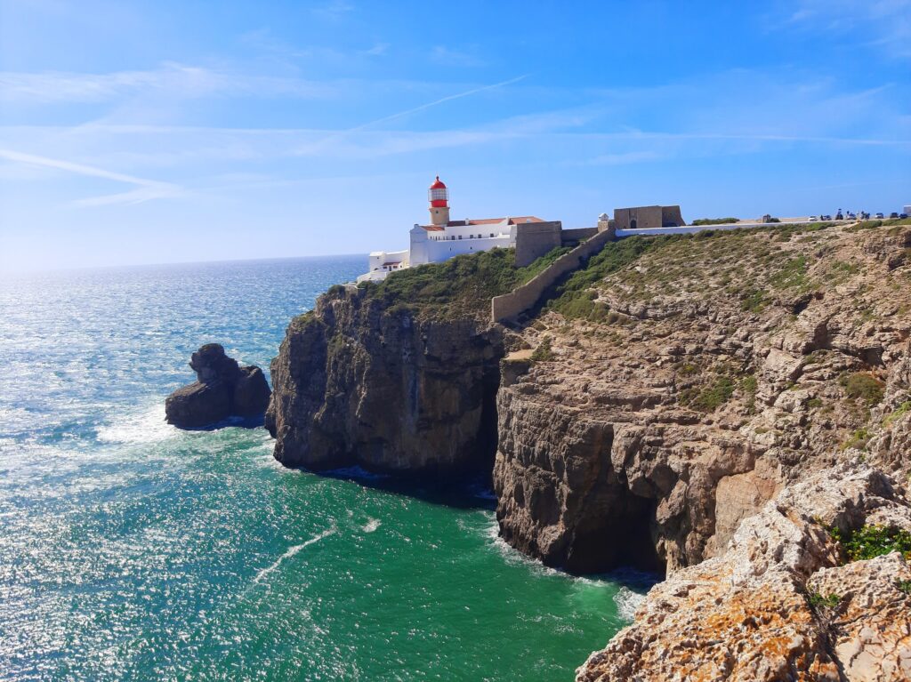 Weißer Leuchtturm mit roter Kuppel auf steiler Felsküste über türkisblauem, sonnenglitzerndem Atlantik.