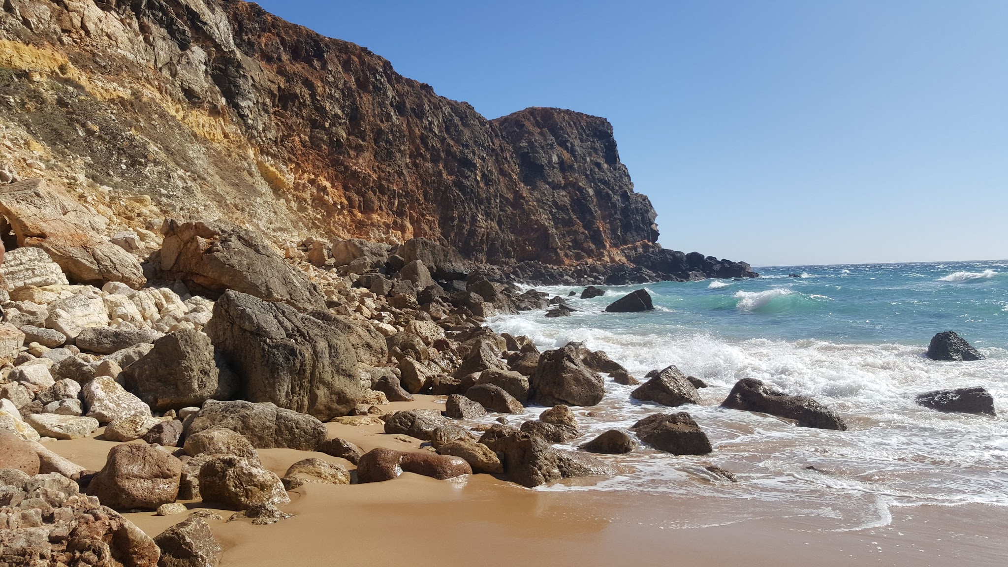 Steile, rötlich-braune Klippe neben felsigem Sandstrand; türkisfarbene Wellen branden unter wolkenlosem Himmel.