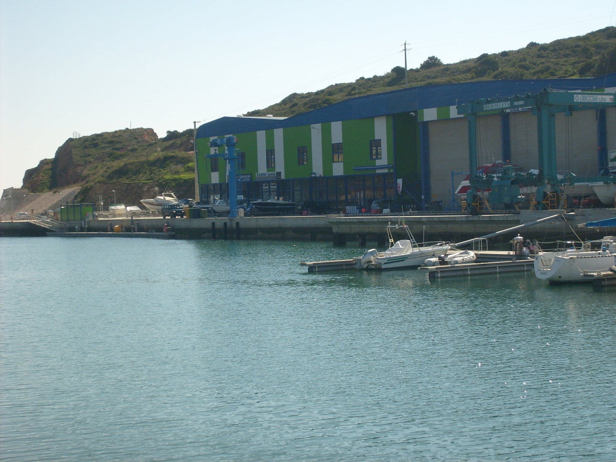 Kleine Marina mit Motorbooten am Steg; dahinter grün-blaues Werftgebäude und bewaldeter Hügel.