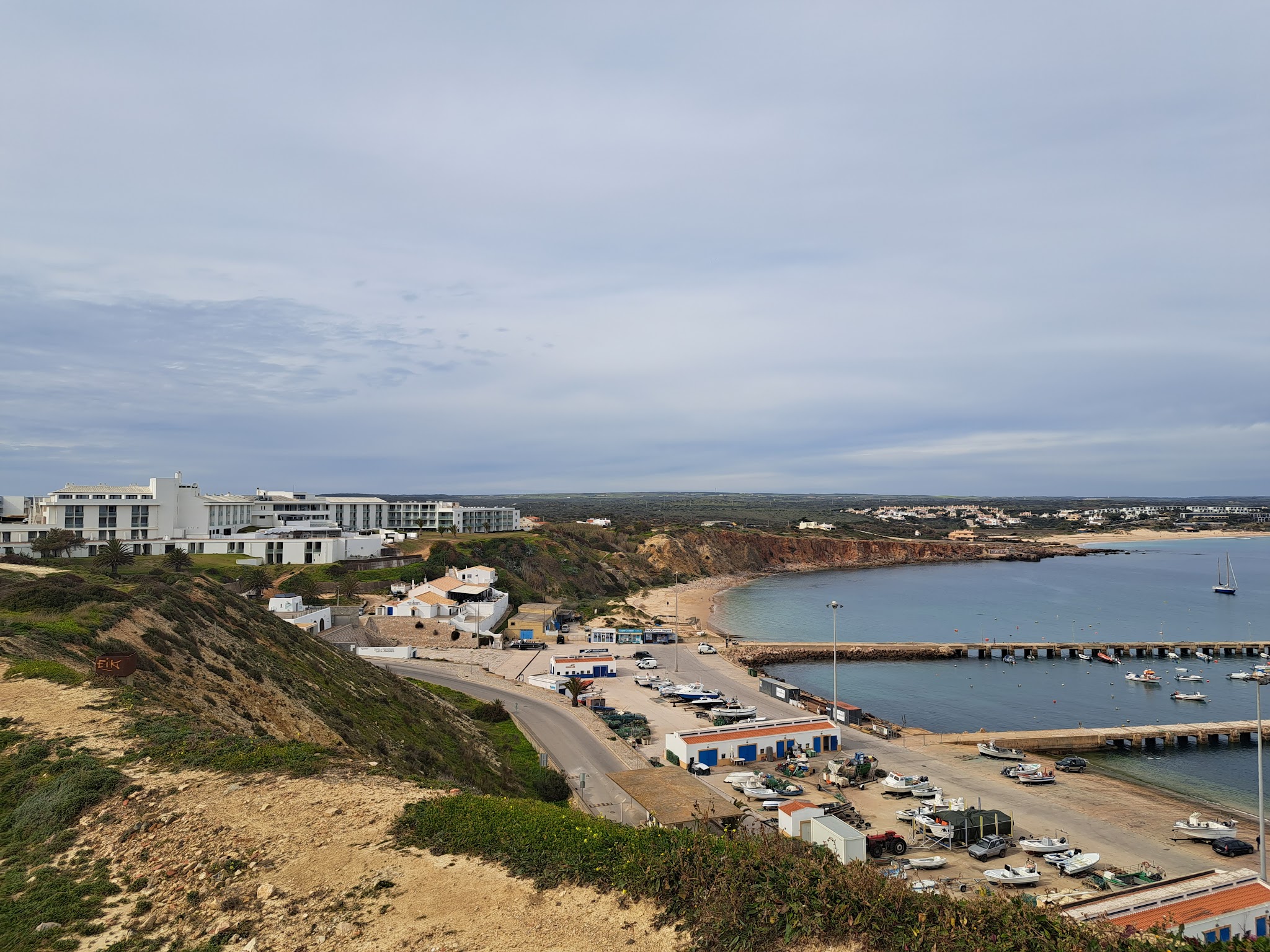 Klippenküste mit weißem Hotel, kleinem Fischerhafen und Booten an ruhiger Bucht unter bewölktem Himmel.