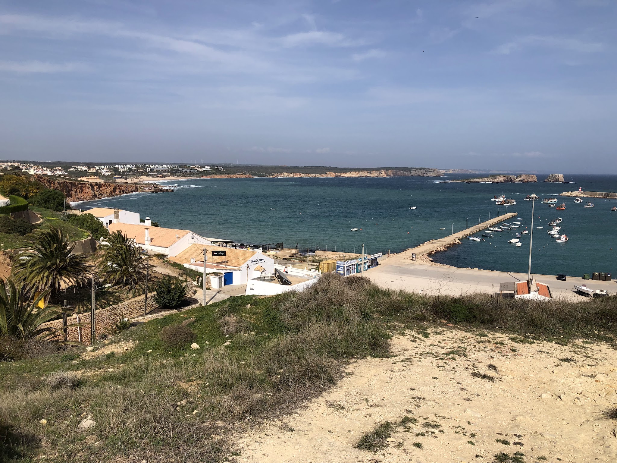 Küstenort mit Palmen, Sandsteinfelsen und langem Pier, an dem kleine Boote im türkisblauen Meer liegen.