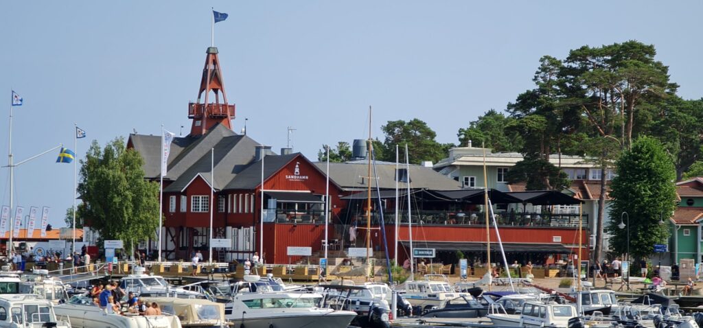 Sommerlicher Jachthafen in Sandhamn; rote Holzbauten, viele Boote und Menschen am Steg.