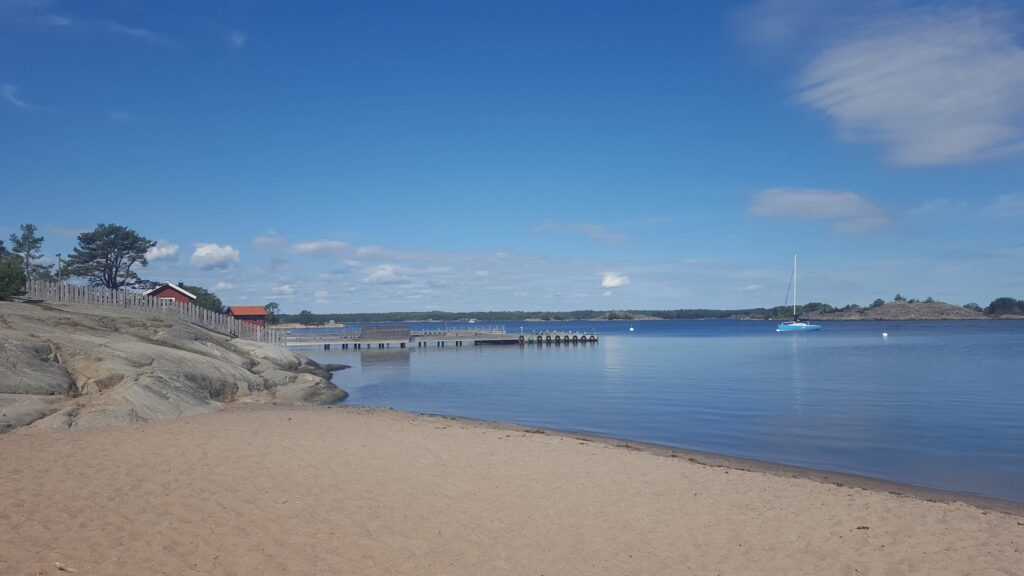 Ruhiger Sandstrand mit Steg; rote Häuschen auf Felsen links, einzelnes blaues Segelboot auf stiller See unter blauem Himmel