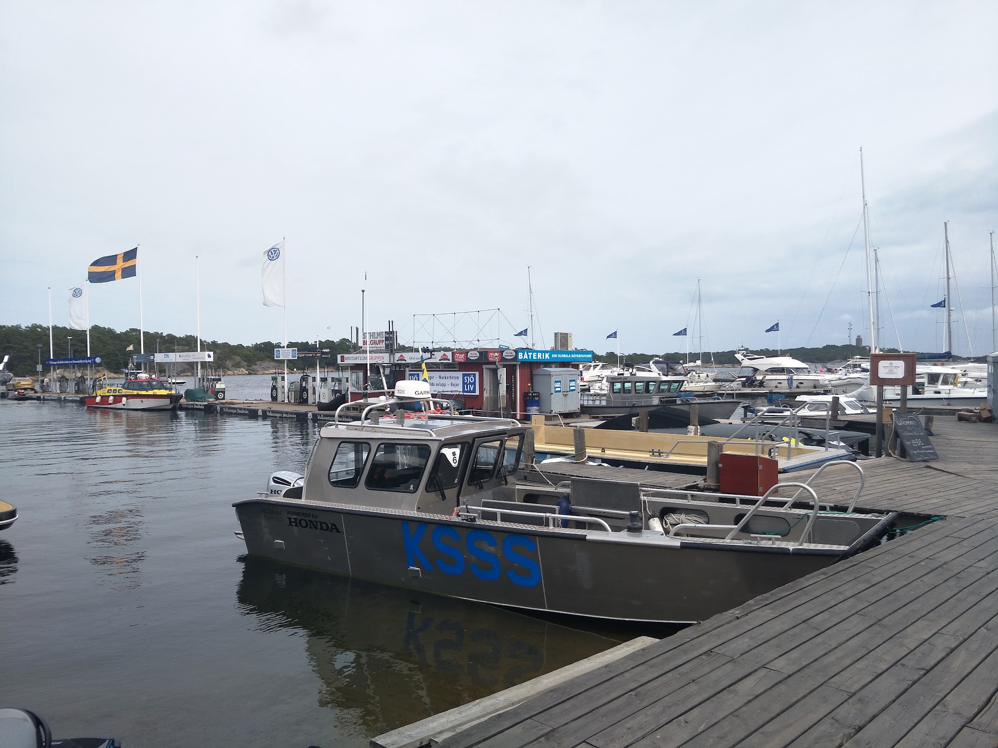 Hafen mit Motor- und Segelbooten, davor graues KSSS-Boot; schwedische Flagge weht am Kai unter bewölktem Himmel