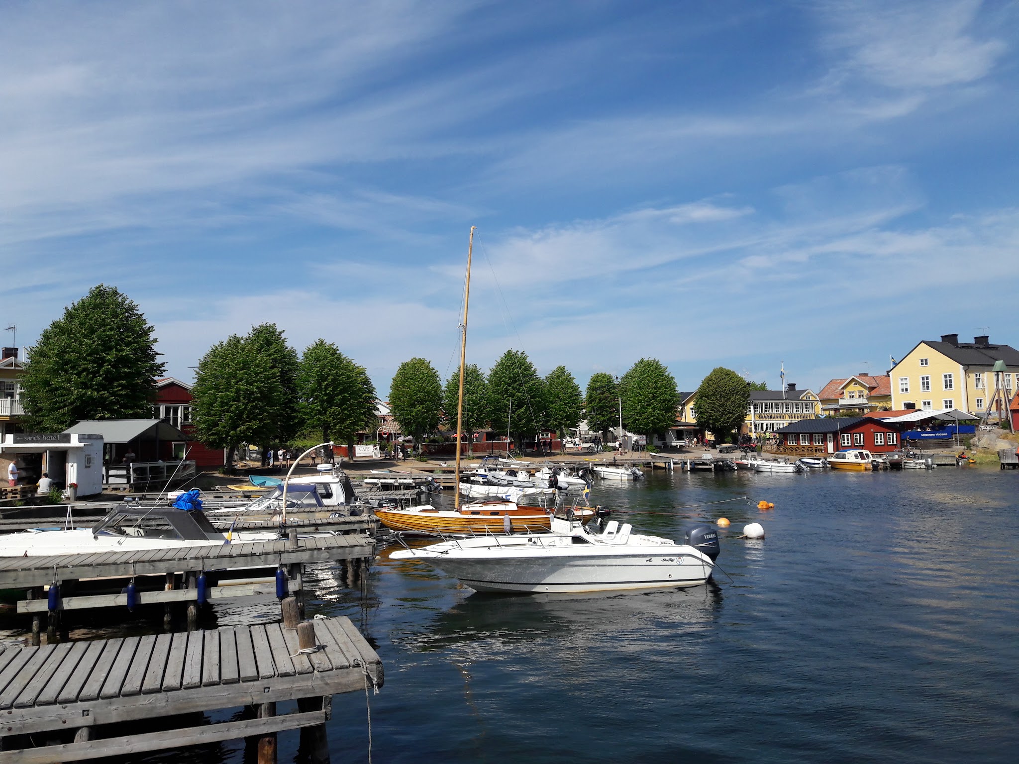 Idyllischer kleiner Hafen mit Booten an Holzstegen, bunte Häuser und grüne Bäume am sonnigen Ufer, blauer Himmel.
