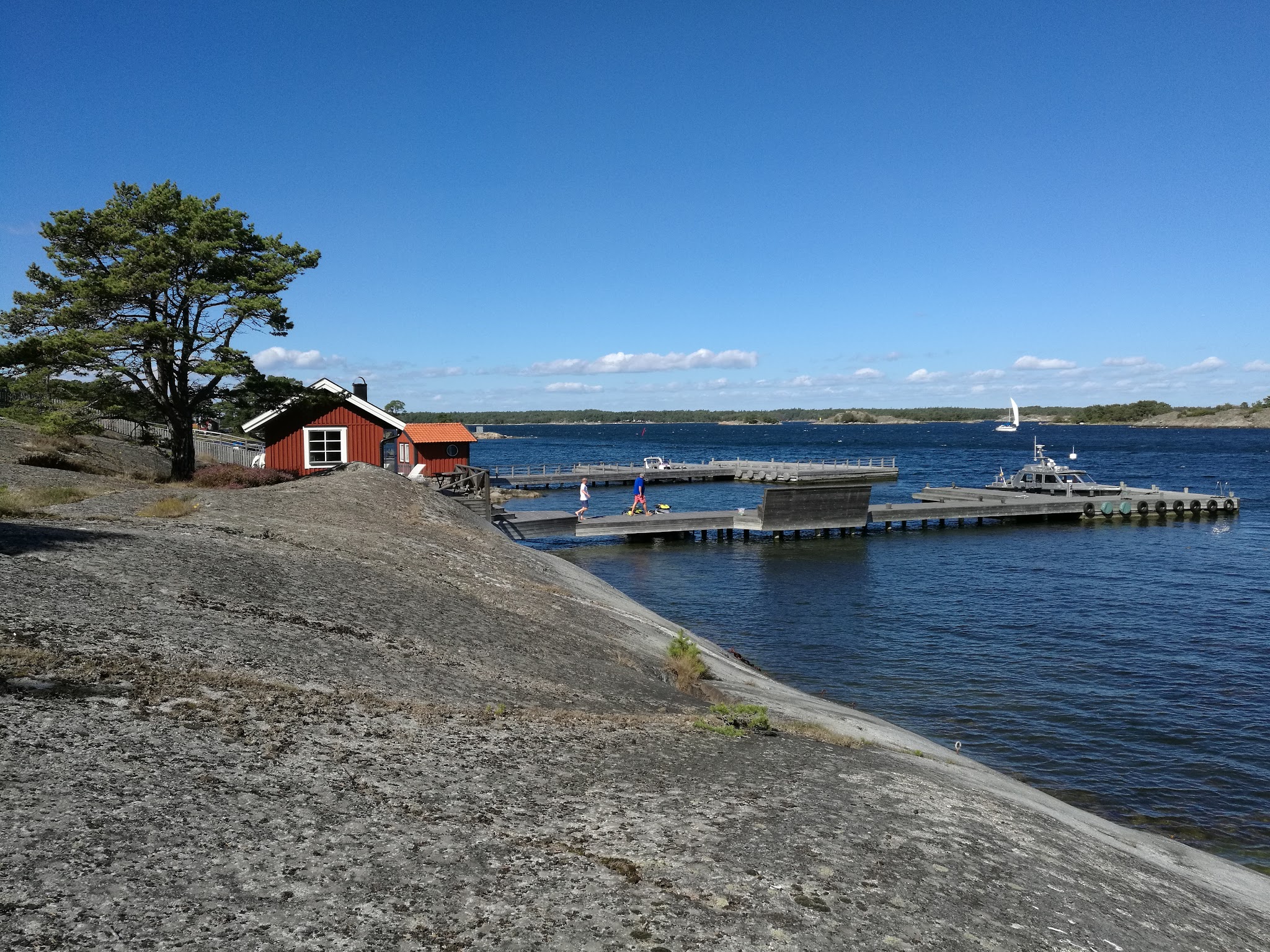 Rote Holzhütte und Pier an felsiger Schärenküste, blaues Meer, Segelboot am Horizont unter klarem Himmel