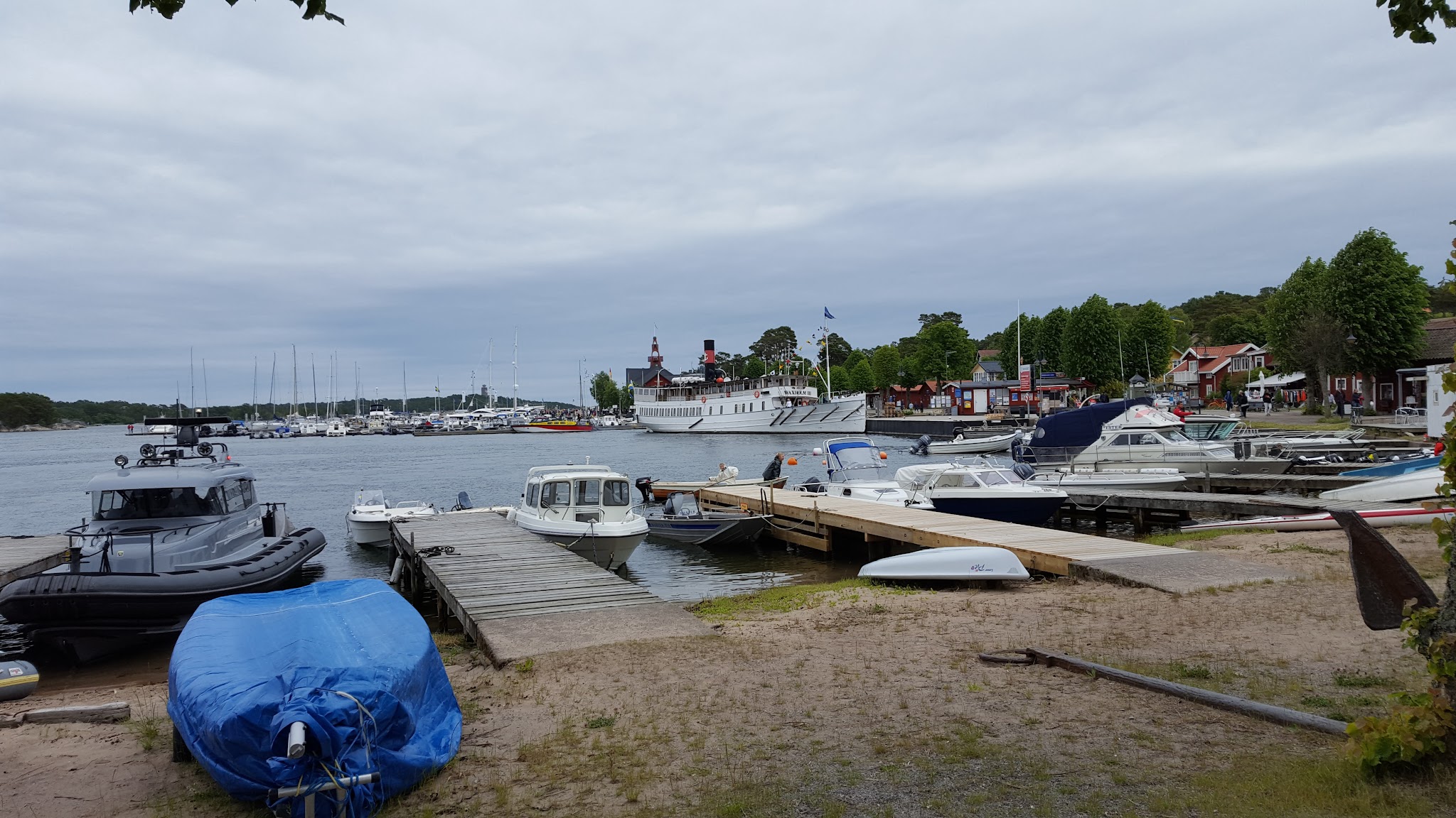 Kleiner Hafen mit Holzstegen; Motorboote, Segelboote und ein großer weißer Dampfer vor bunten Uferhäuschen.