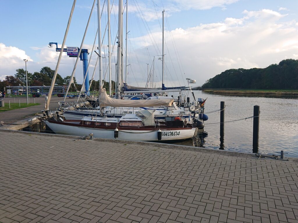 Mehrere Segelboote liegen vertäut an einem ruhigen Hafenbecken; Pflasterkai im Vordergrund, Wald am Horizont.