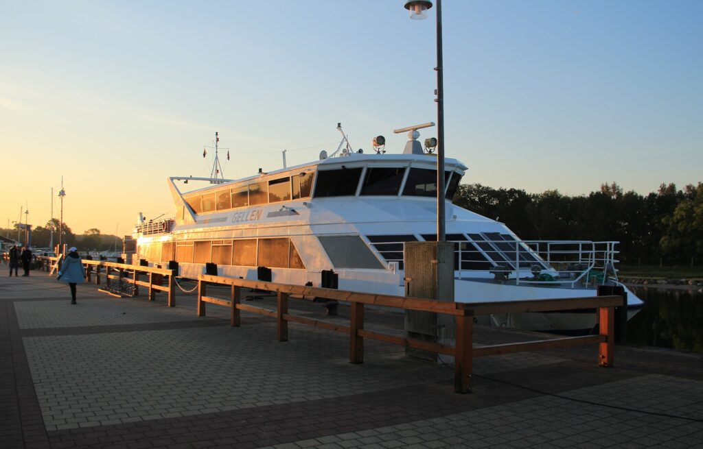 Katamaranfähre „Gellen“ liegt bei Sonnenuntergang am Anleger; vereinzelte Passanten gehen über die Promenade.