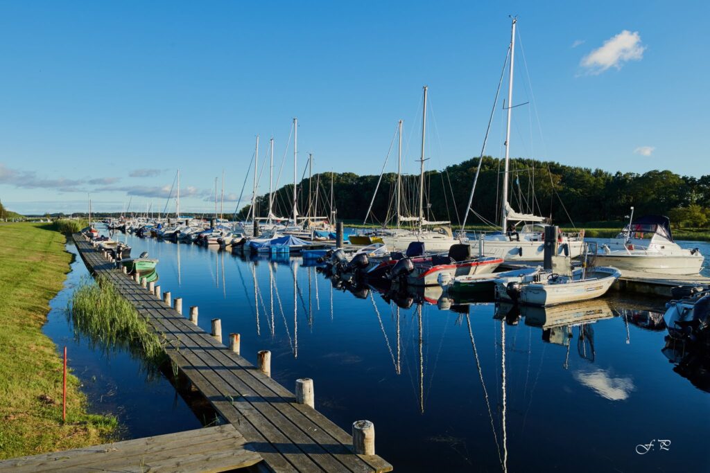 Holzsteg entlang ruhigem Hafen; zahlreiche Segel- und Motorboote spiegeln sich im klaren Wasser unter blauem Himmel.