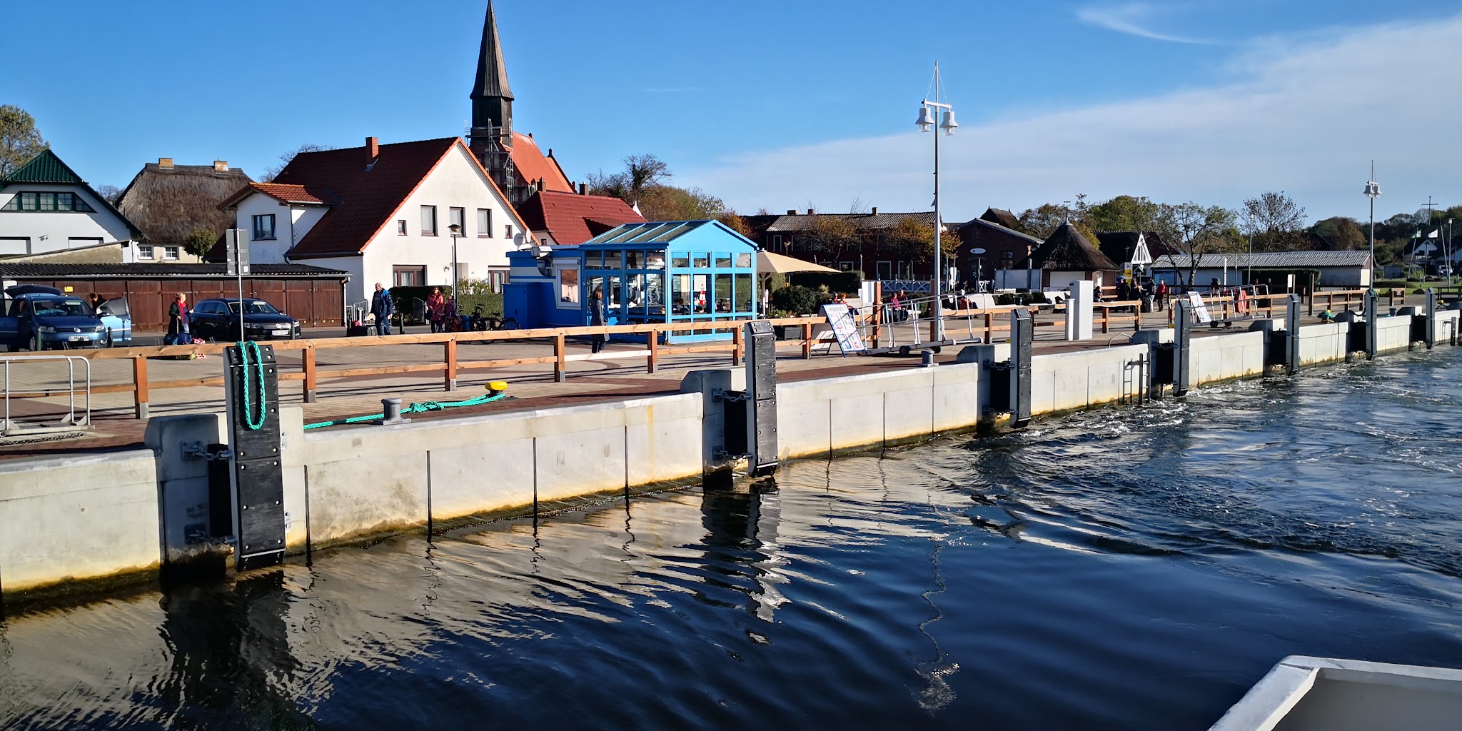 Hafenpromenade mit Betonpier, blauem Glaspavillon und Dorfhäusern; Kirche ragt im Hintergrund, Wasser im Vordergrund
