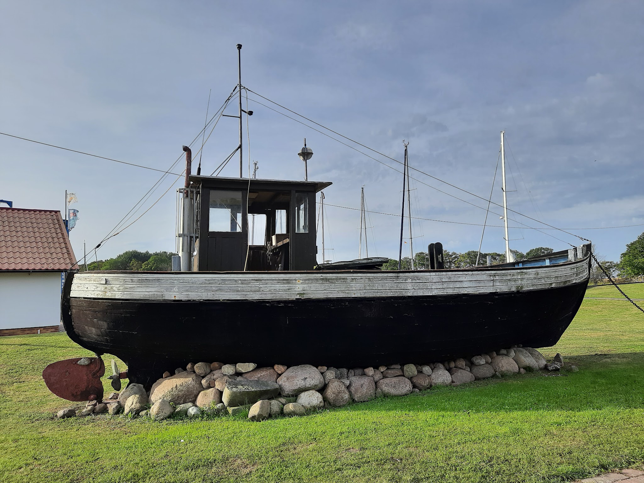 Altes schwarzes Holzfischerboot steht auf Steinhaufen im Gras, Masten und Seile vor blauem Himmel.