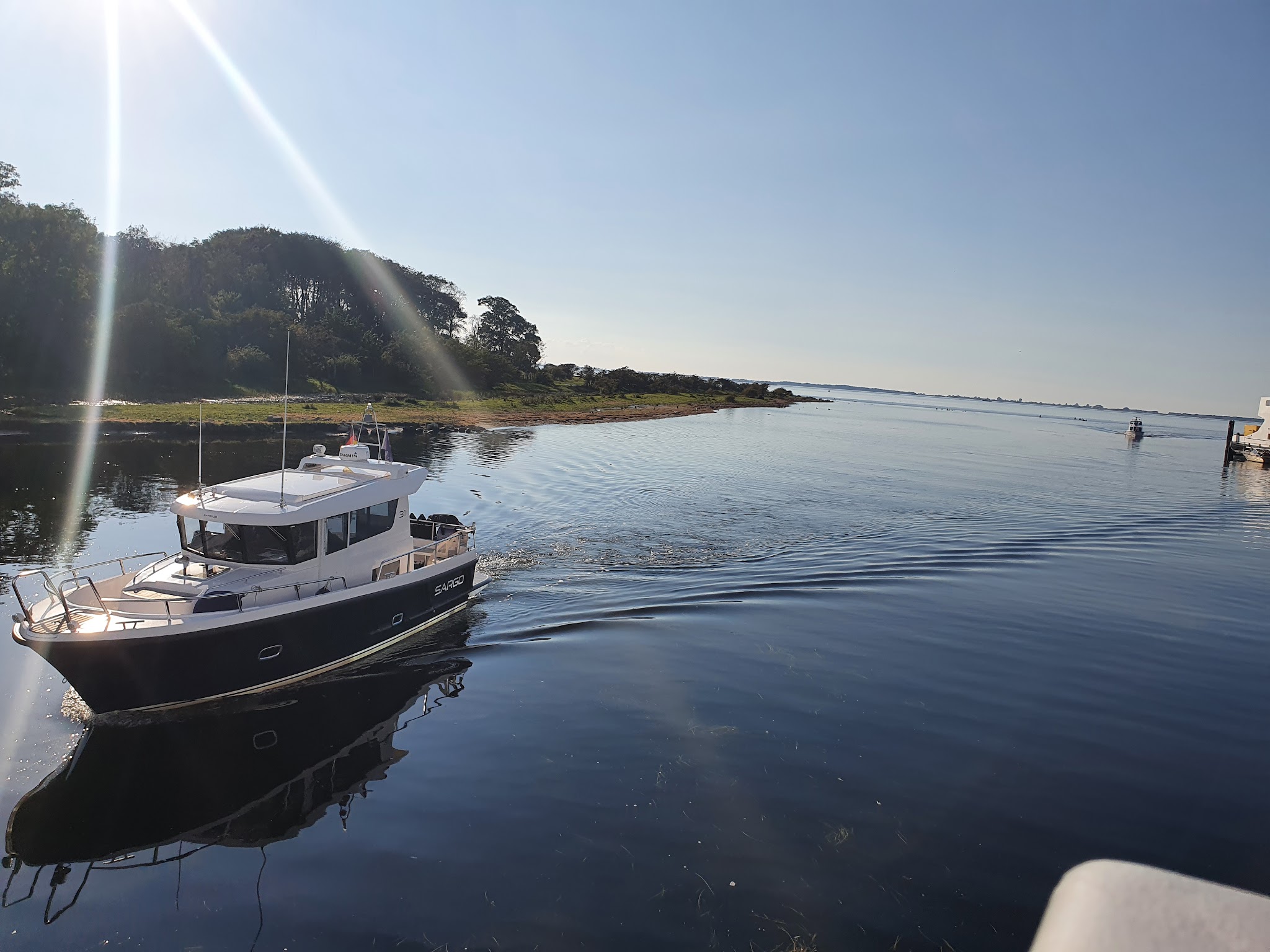 Kleines Motorboot zieht Spur auf ruhigem, sonnendurchflutetem Fluss nahe bewaldetem Ufer.