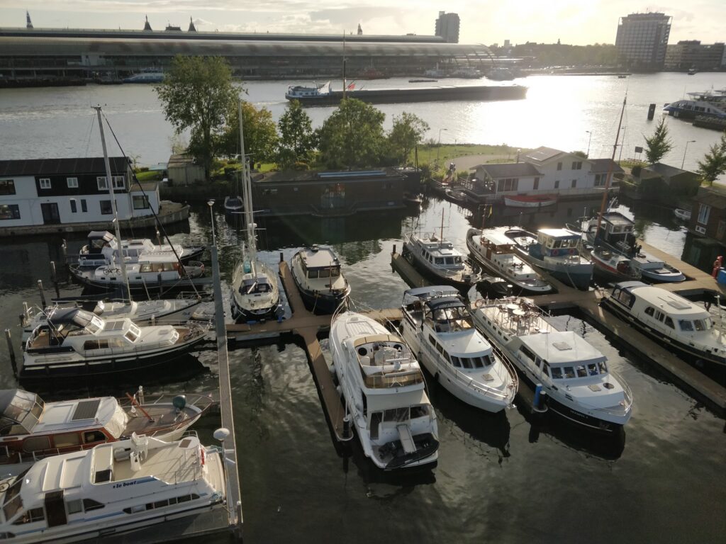 Mehrere Yachten liegen in einer kleinen Marina; Sonnenlicht glitzert auf dem Wasser, Stadtgebäude im Hintergrund.
