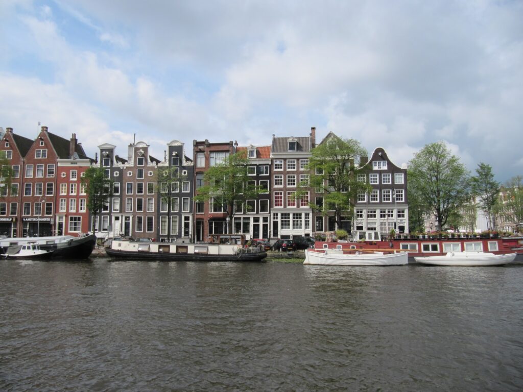 Schmale Giebelhäuser am Amsterdamer Kanal, davor festgemachte Hausboote auf ruhigem Wasser unter leicht bewölktem Himmel
