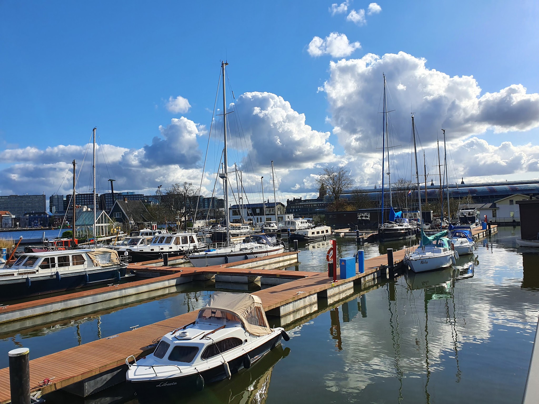 Kleiner Jachthafen mit Segel- und Motorbooten an Holzstegen, ruhiges Wasser spiegelt Wolken am sonnigen Himmel.