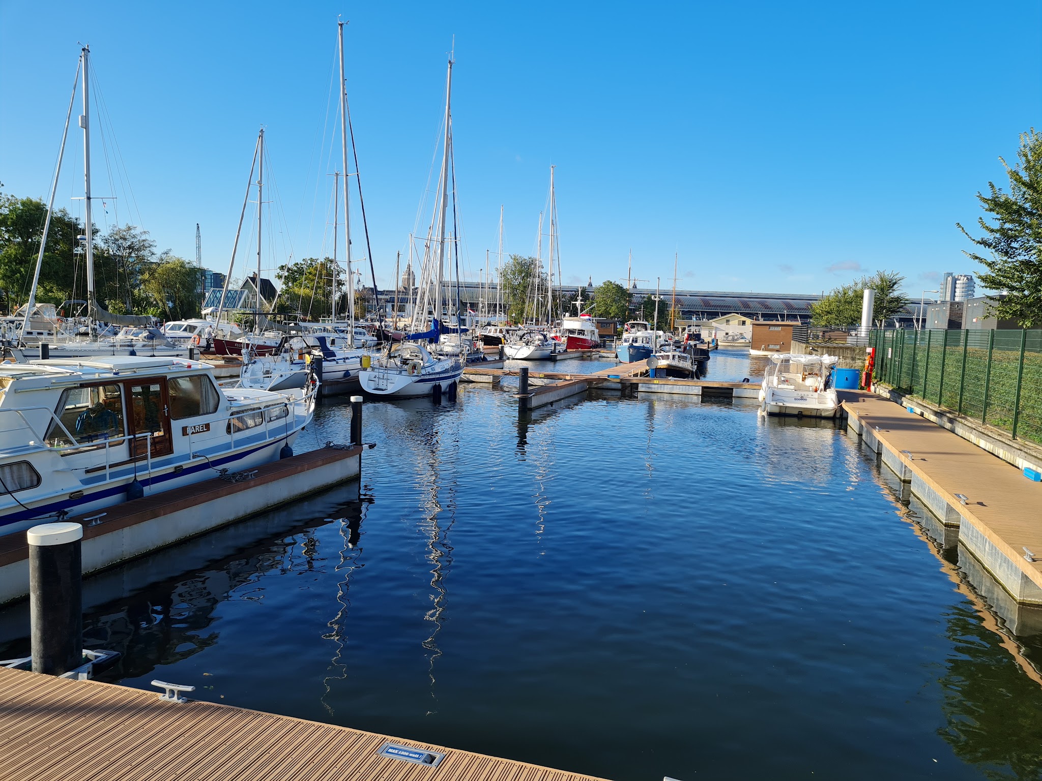 Ruhiger Yachthafen mit zahlreichen Segelbooten an Stegen, spiegelndes Wasser unter klarem blauem Himmel.
