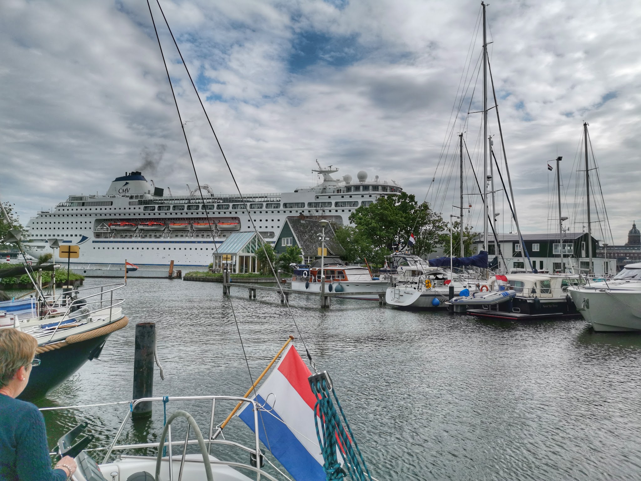 Kreuzfahrtschiff passiert kleinen Jachthafen mit Segelbooten; niederländische Flagge im Vordergrund