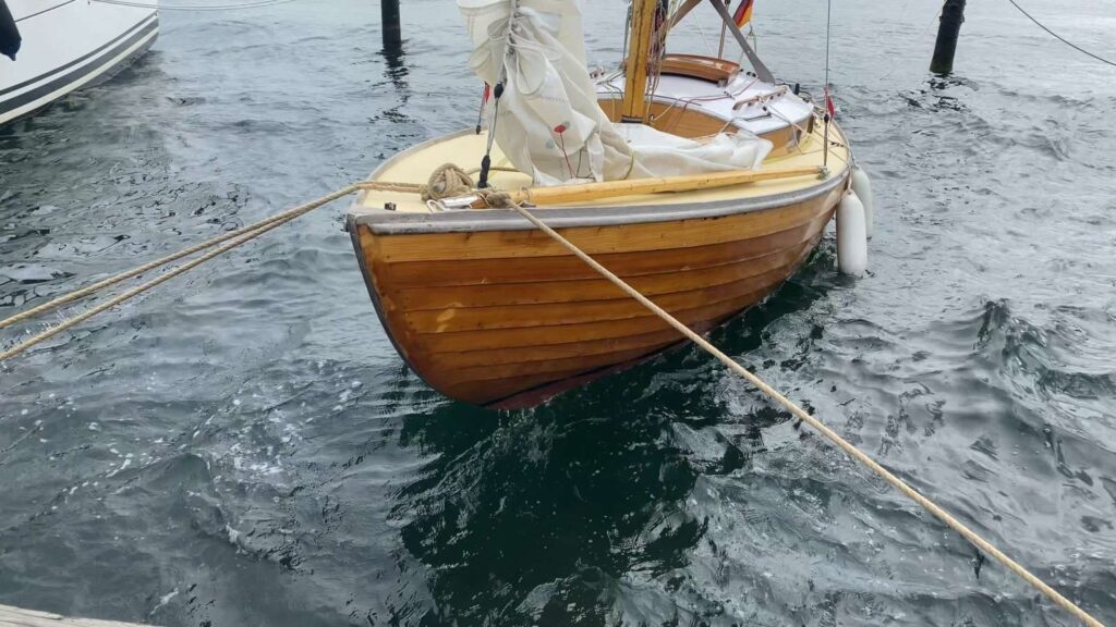 Kleines hölzernes Segelboot mit eingepacktem Segel, mit Tauen am Steg vertäut, auf leicht bewegter Wasseroberfläche.