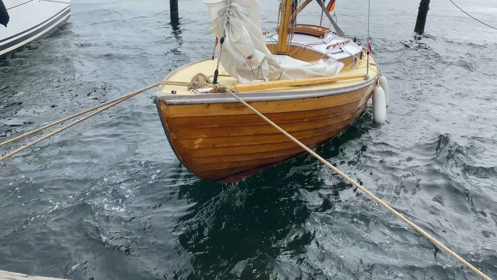Kleines hölzernes Segelboot mit eingepacktem Segel, mit Tauen am Steg vertäut, auf leicht bewegter Wasseroberfläche.
