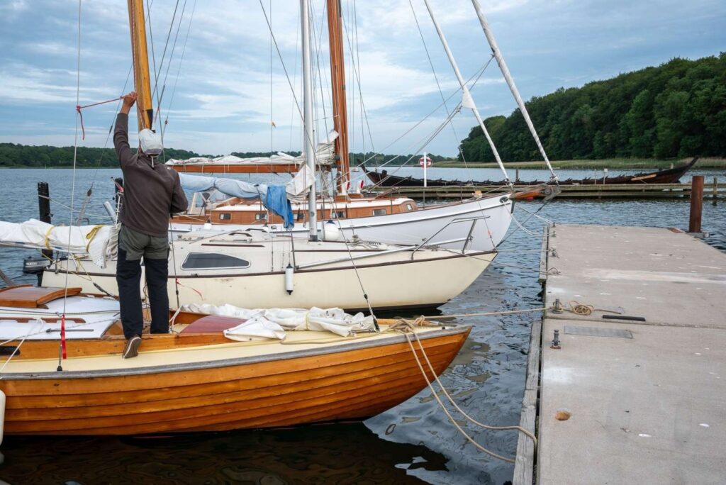 Segler spannt eine Leine an einem Holz-Segelboot am Steg; weitere Boote liegen im ruhigen Hafen vor Waldkulisse.