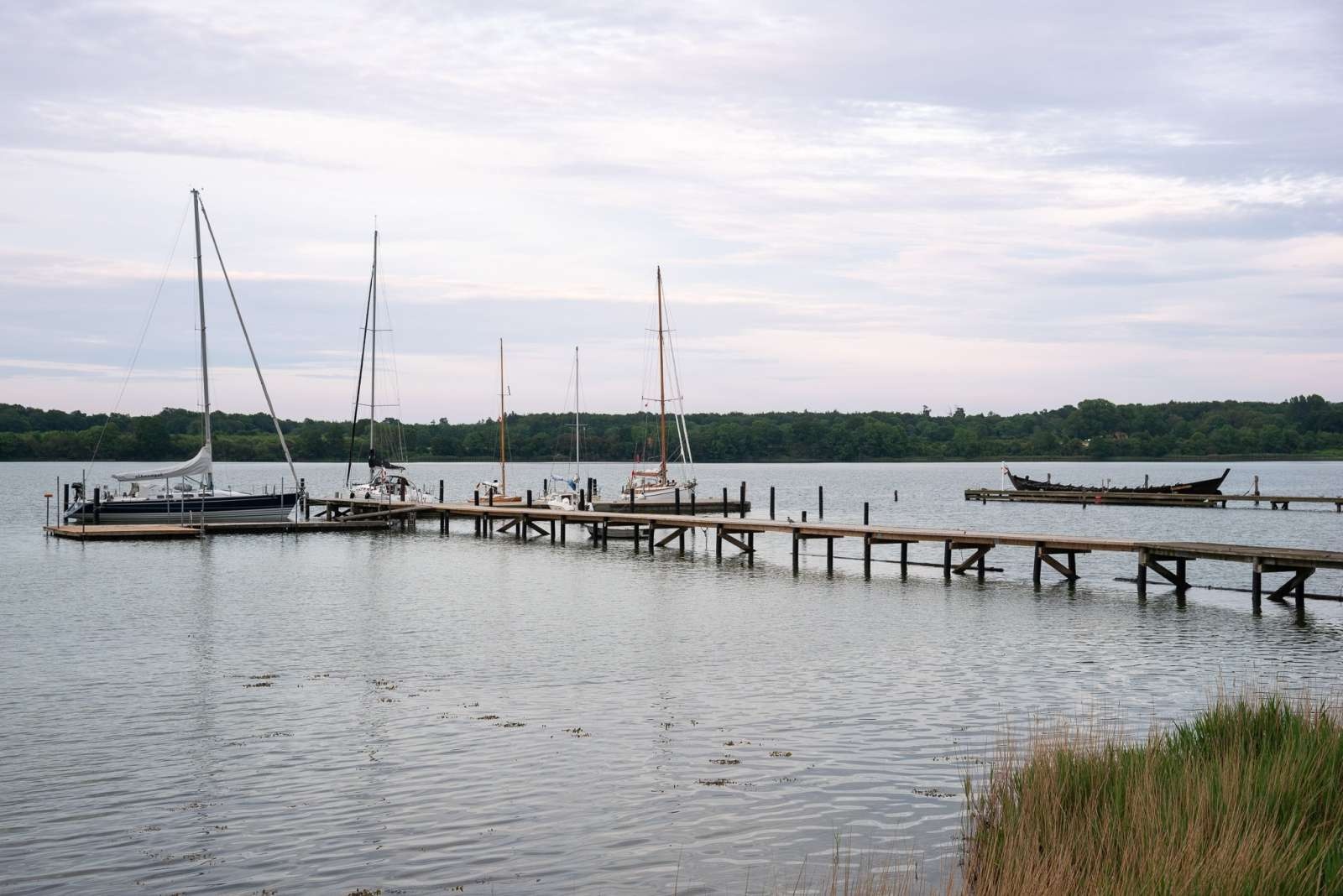 Holzsteg ragt in ruhigen See; mehrere Segelboote und ein langes Holzboot liegen vor bewaldetem Ufer unter Wolken.