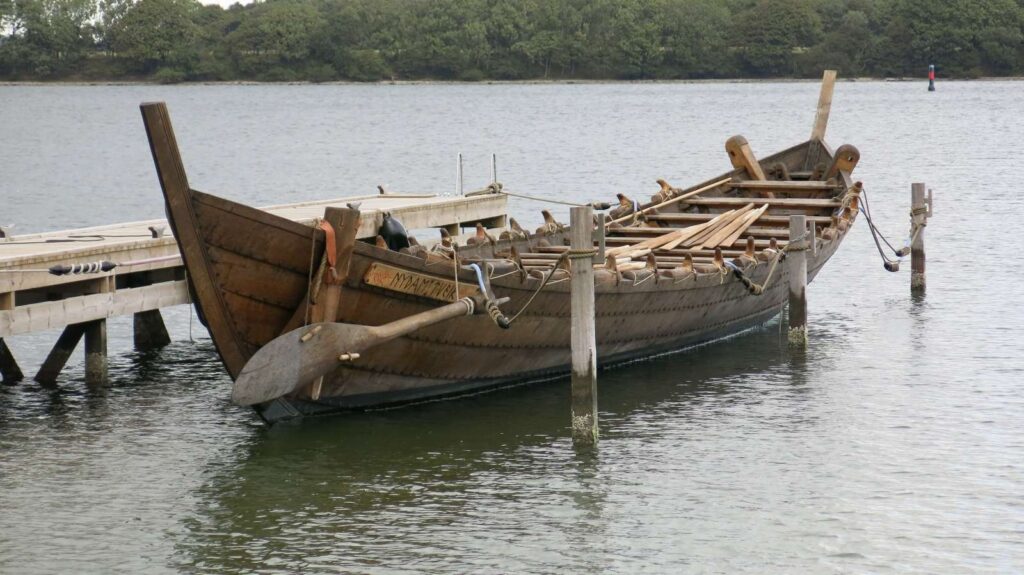 Historisches Holzschiff mit losen Rudern vertäut an kleinem Steg auf stiller Wasserfläche.
