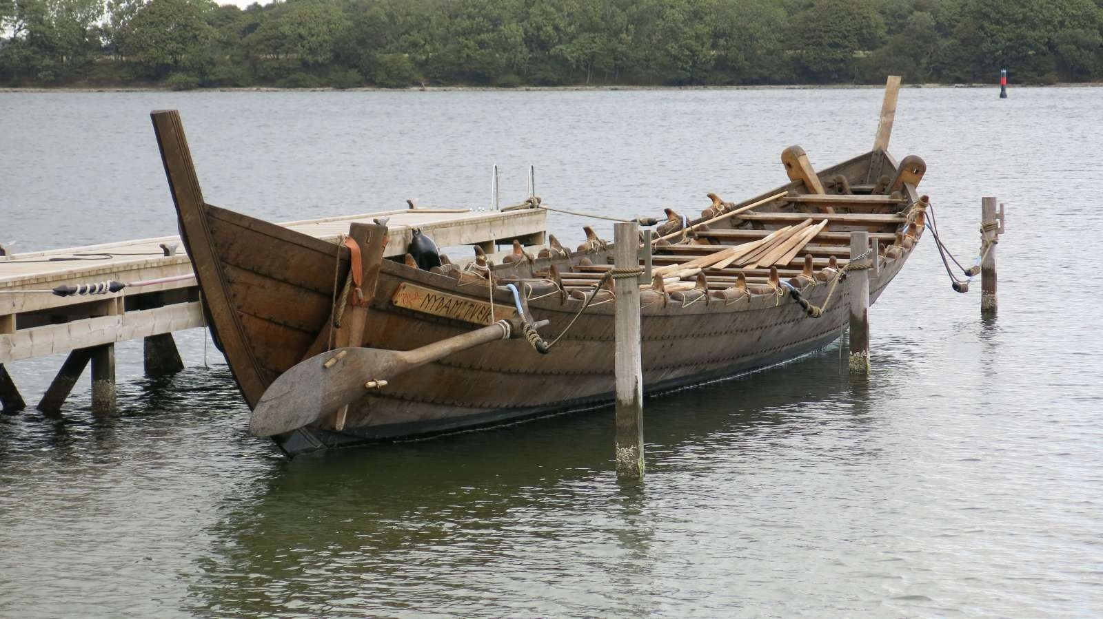Historisches Holzschiff mit losen Rudern vertäut an kleinem Steg auf stiller Wasserfläche.