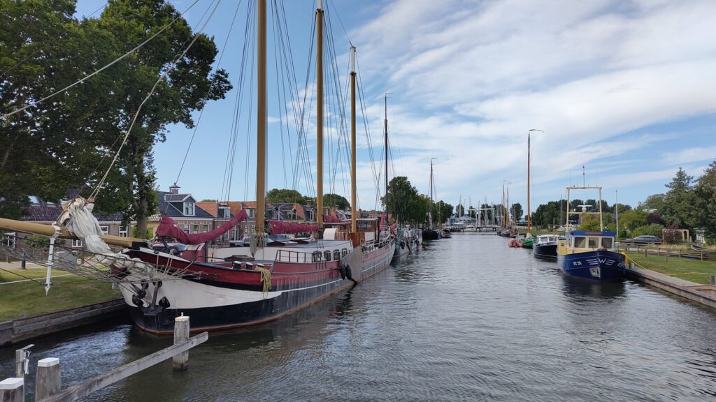 Mehrmastige Segelschiffe liegen an einem ruhigen Kanal vor Backsteinhäusern und Bäumen unter leicht bewölktem Himmel.