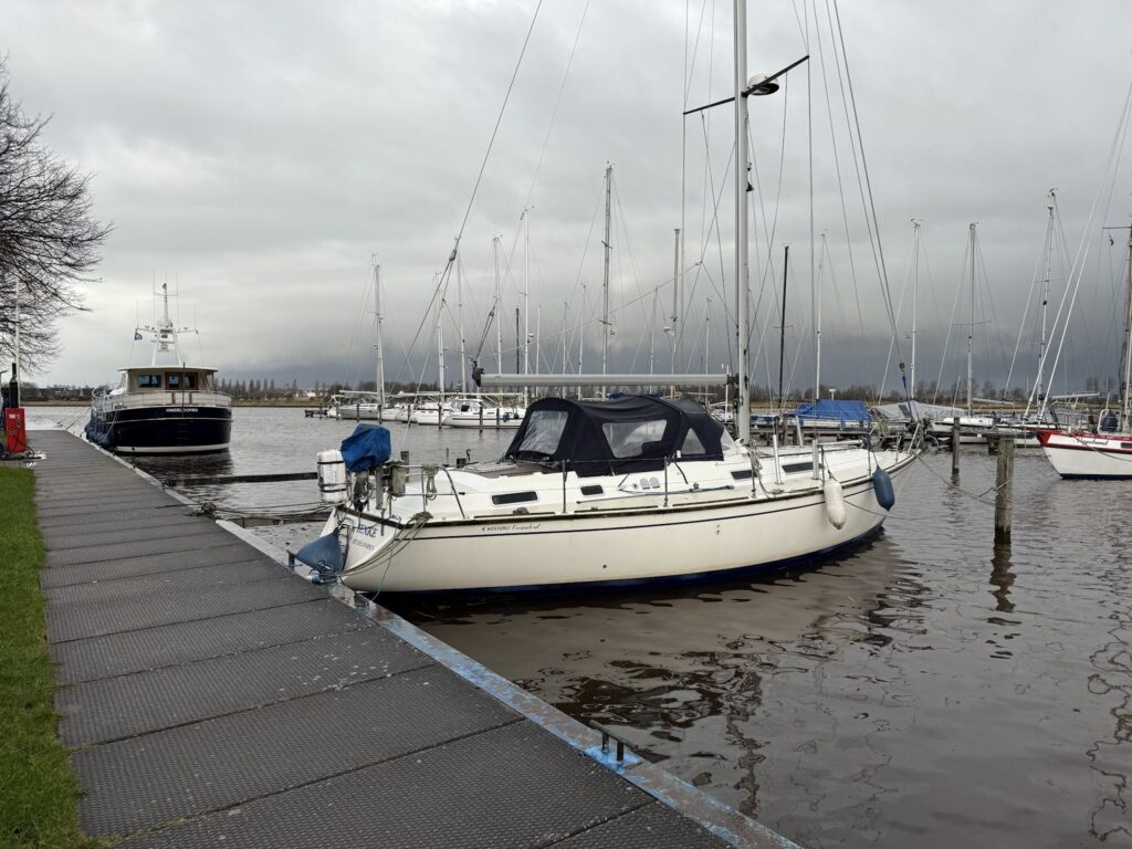 Weißes Segelboot liegt an Steg im Hafen; weitere Boote, Masten und grauer, wolkenverhangener Himmel im Hintergrund