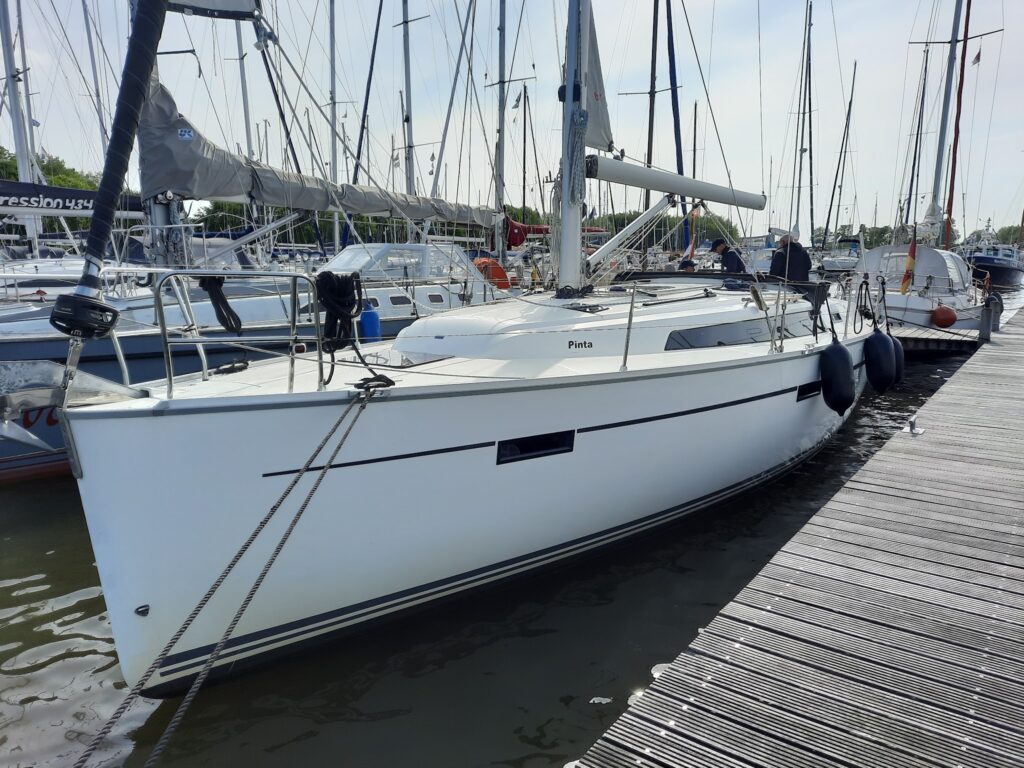 Weiße Segelyacht „Pinta“ im Hafen, an Steg vertäut; zwei Personen an Deck, ringsum Masten vieler Boote