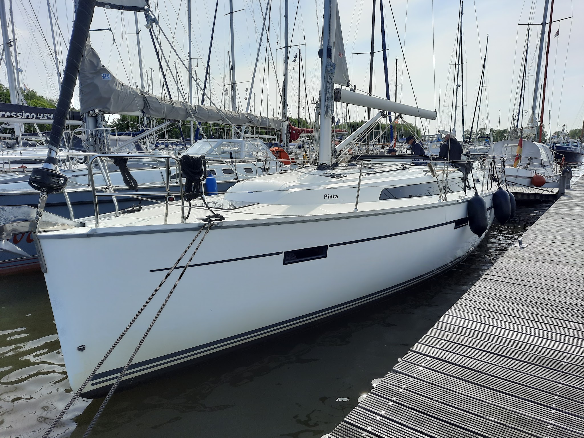 Weiße Segelyacht „Pinta“ im Hafen, an Steg vertäut; zwei Personen an Deck, ringsum Masten vieler Boote