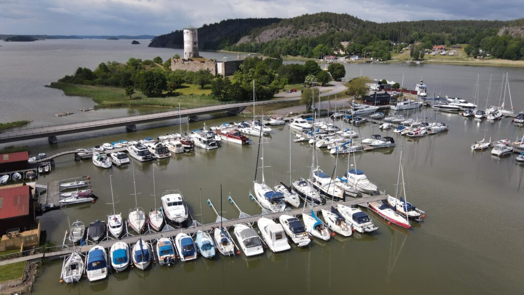 Yachthafen voller Segel- und Motorboote; dahinter eine Insel mit mittelalterlichem Burgturm, umgeben von Wasser und Wald.
