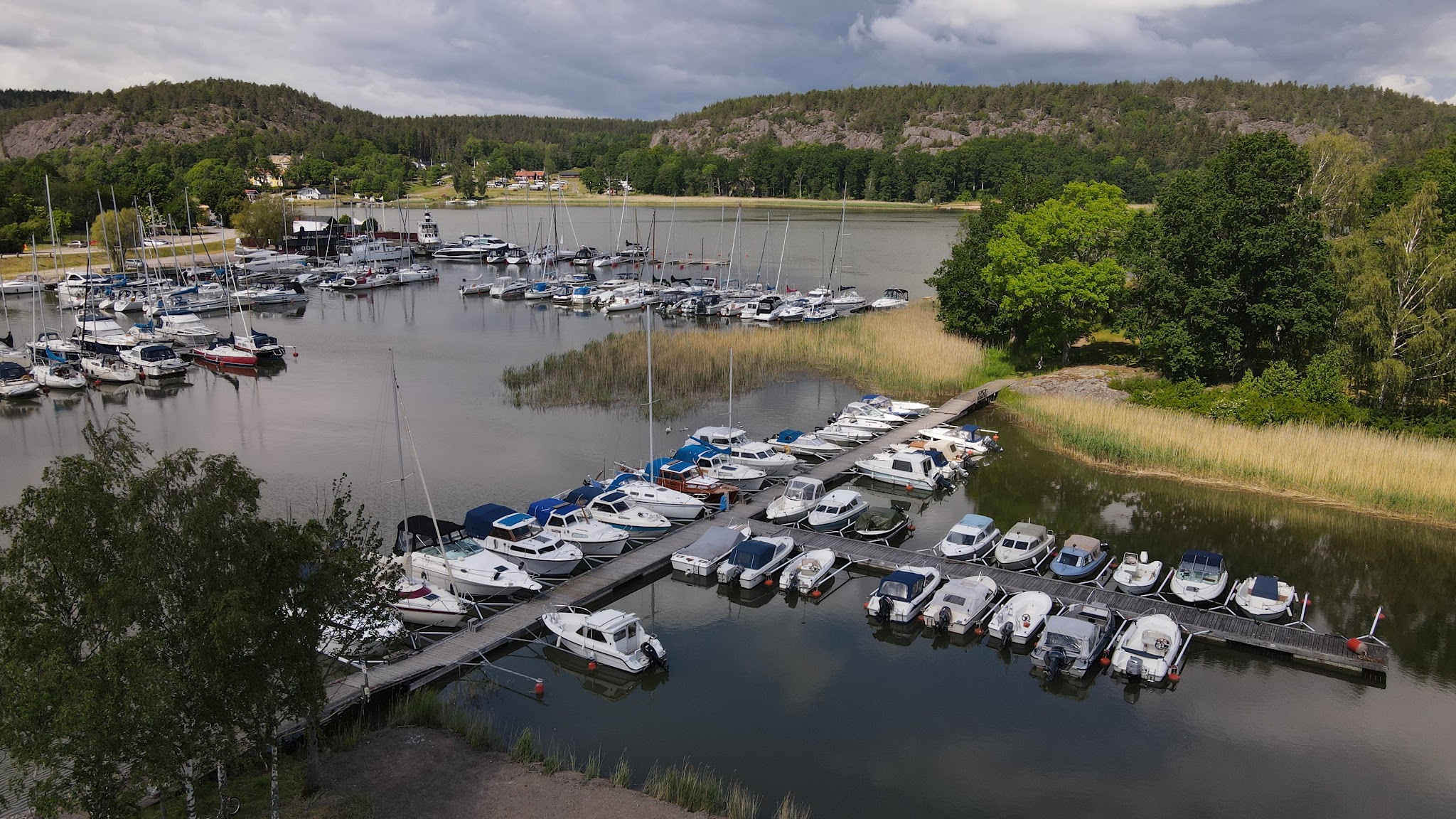 Ruhiger Yachthafen; Reihen weißer Motor- und Segelboote an Stegen, umgeben von Waldhügeln unter wolkigem Himmel.