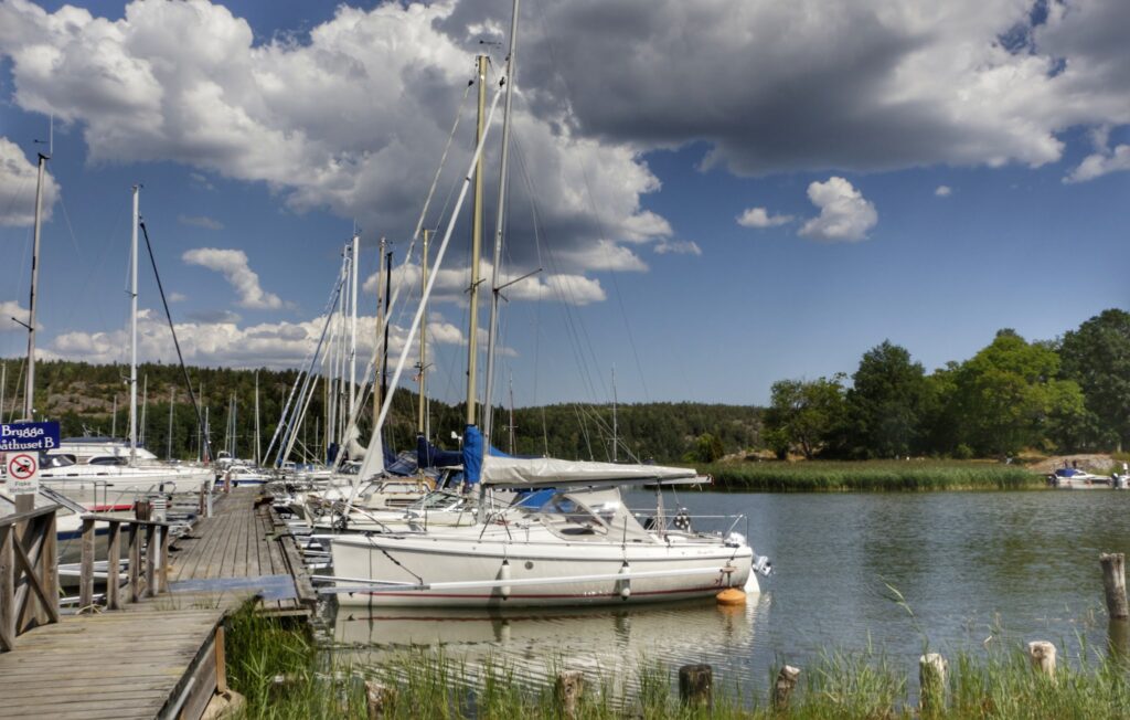 Mehrere Segelboote liegen an einem Holzsteg in ruhigem Hafen, Waldkulisse und Wolken am Himmel.