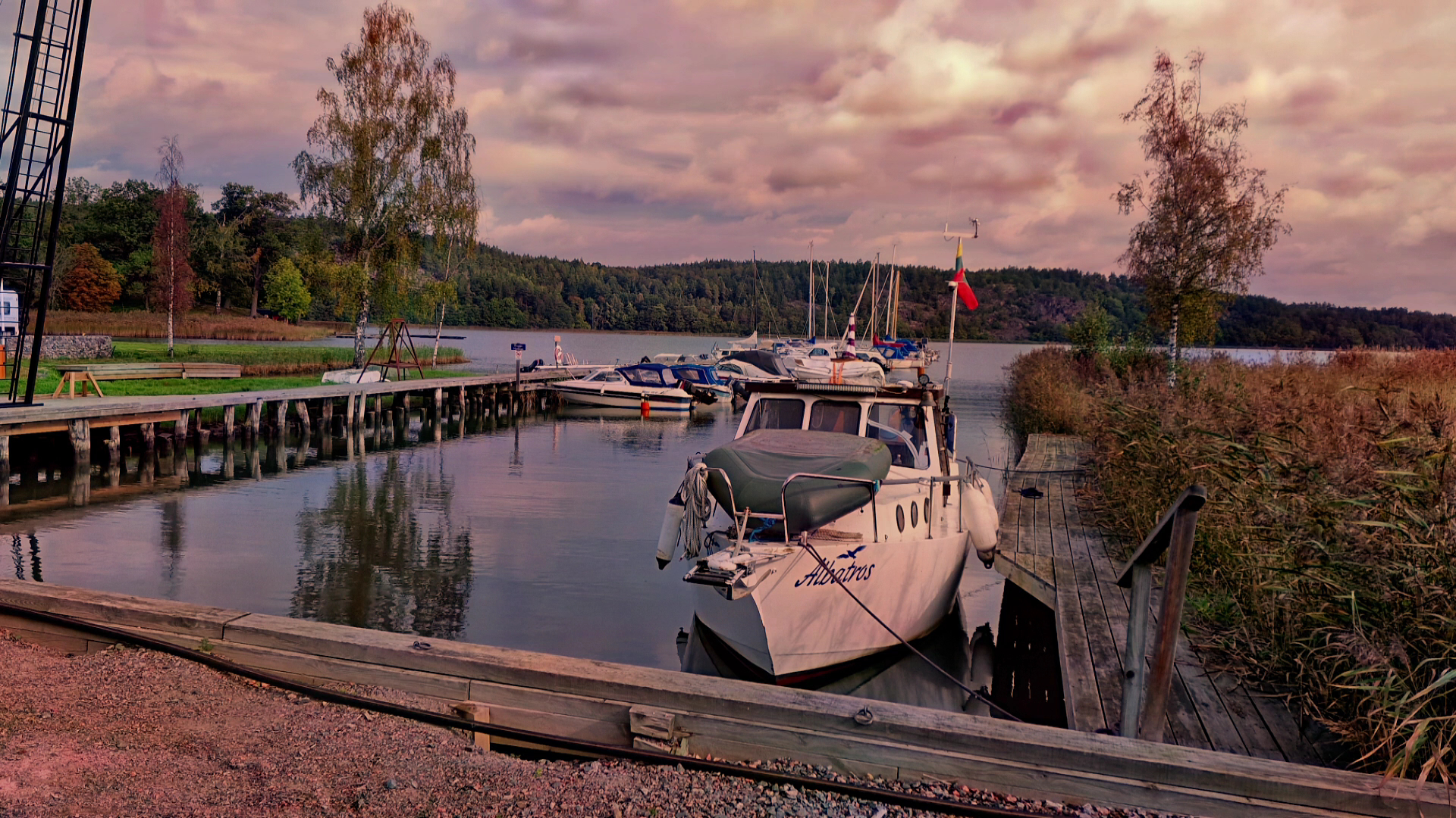Kleine Marina; mehrere Boote liegen am Holzsteg vor bewaldetem Ufer unter rosa Abendhimmel.