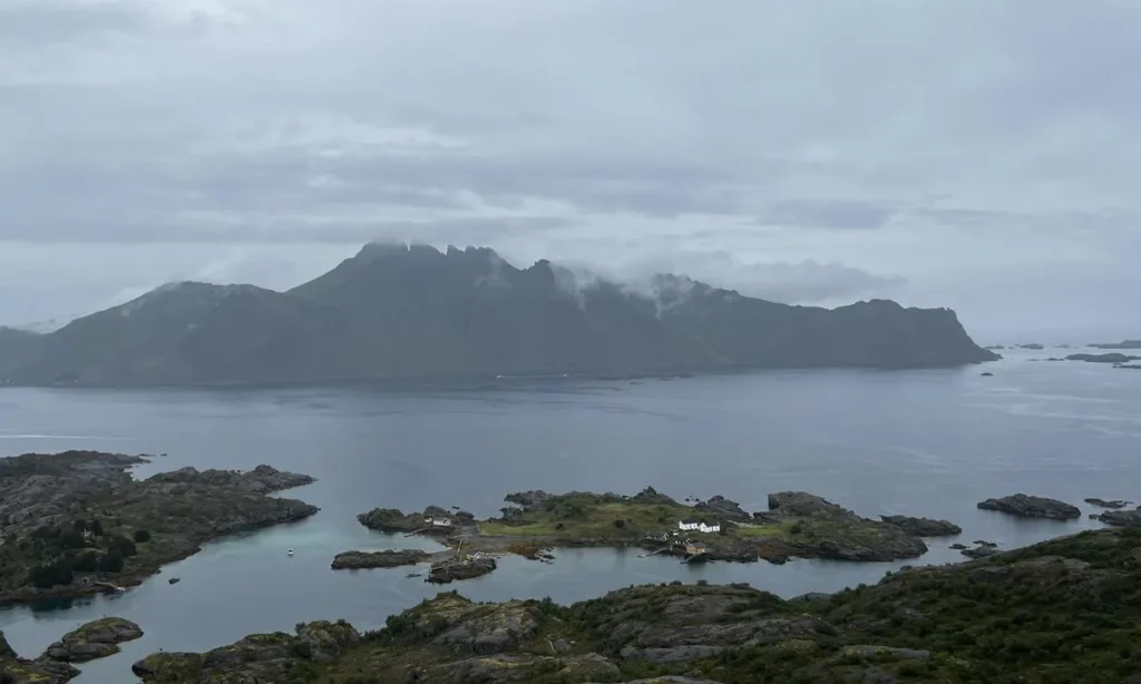 Grauer, wolkenverhangener Fjord mit hohen Bergsilhouetten; vorgelagerte Schäreninsel mit wenigen weißen Häusern.