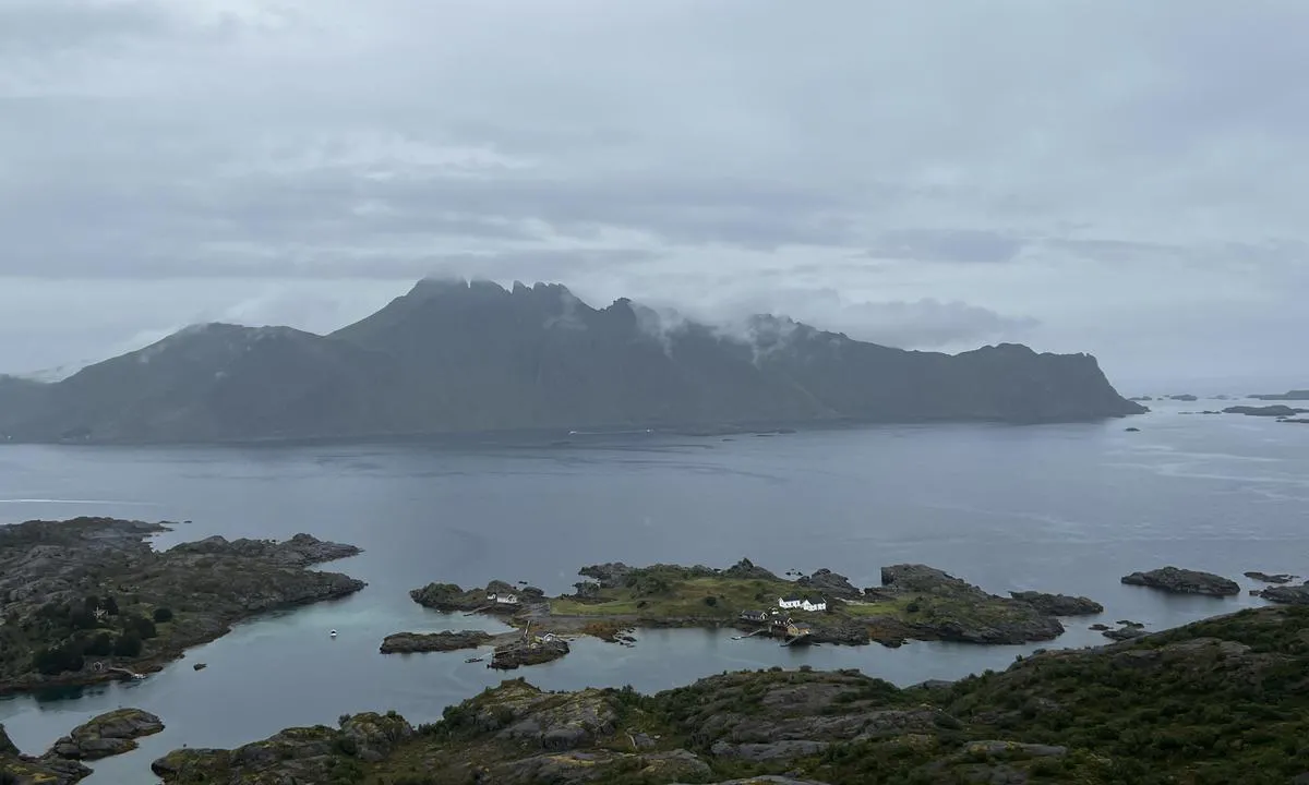Grauer, wolkenverhangener Fjord mit hohen Bergsilhouetten; vorgelagerte Schäreninsel mit wenigen weißen Häusern.