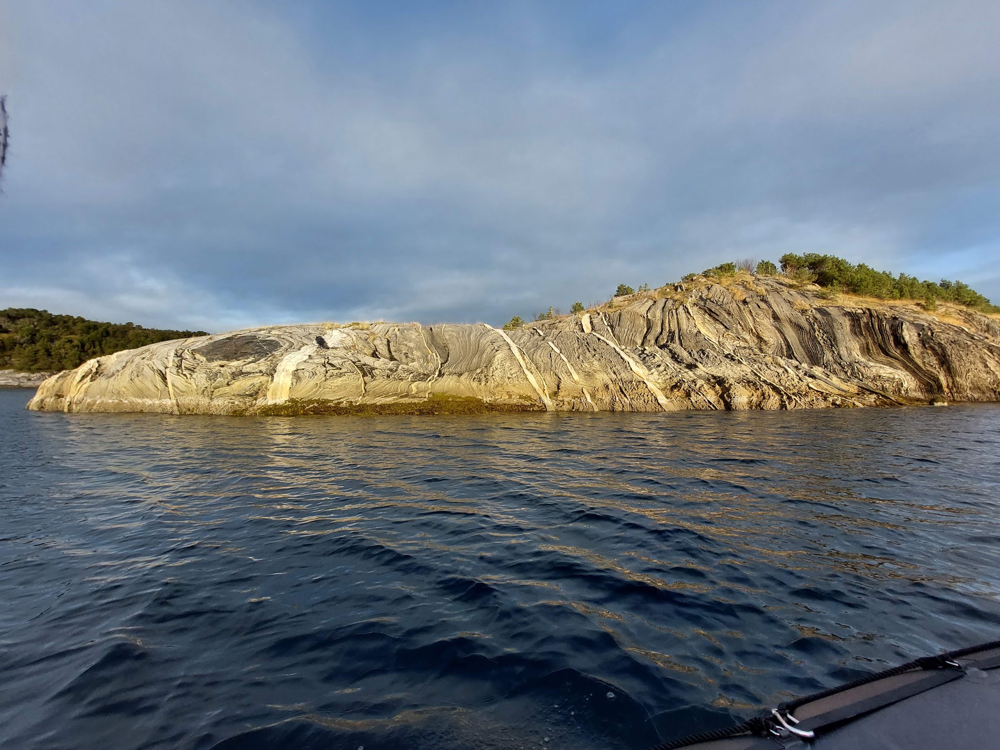 Welliges Meer vor kahl gestreifter Felsinsel mit etwas Wald, beleuchtet von tiefem Licht unter teils bewölktem Himmel